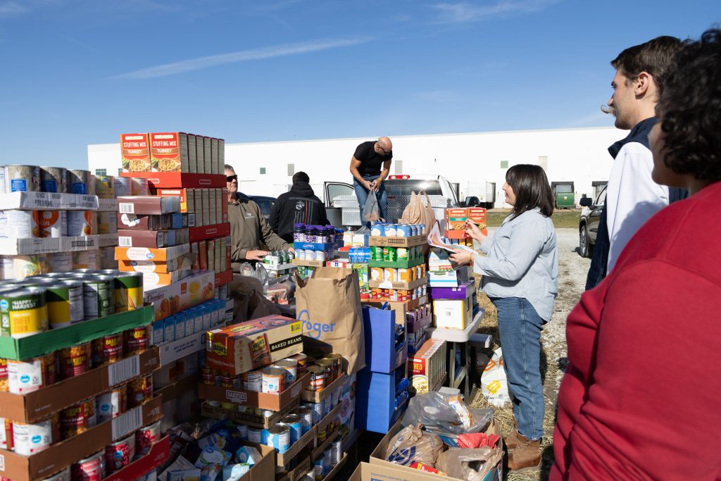Volunteers sort and hand out canned goods at an outdoor community food drive, with stacked boxes and bags nearby.