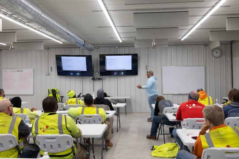 Group of workers in high-visibility vests seated at tables watching a presenter in a light blue shirt in a workshop or training room with screens on the wall
