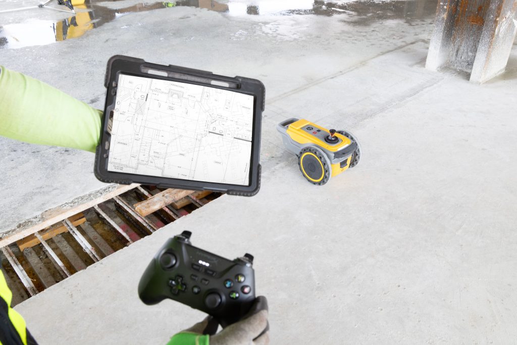 Construction worker holds a rugged tablet displaying a blueprint over a concrete site with a trench nearby and a small yellow device on the ground nearby.