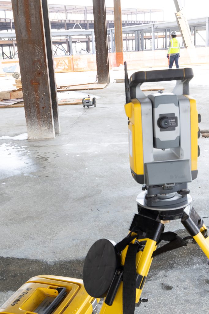 Construction site with a yellow surveying instrument on a tripod in the foreground and a worker in a high‑visibility vest in the background.