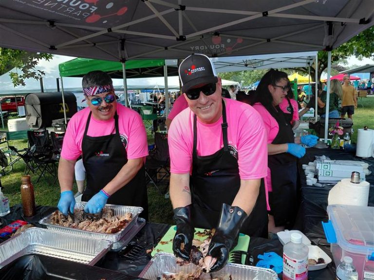 Two volunteers in pink shirts and black aprons cut smoked meat at an outdoor BBQ booth under a canopy outside.