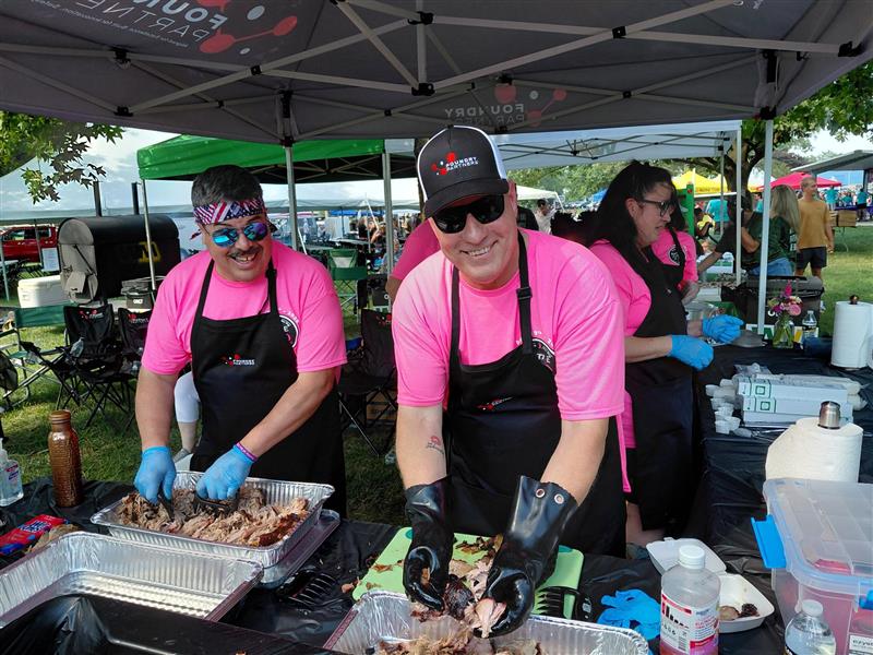Two volunteers in pink shirts and black aprons cut smoked meat at an outdoor BBQ booth under a canopy outside.