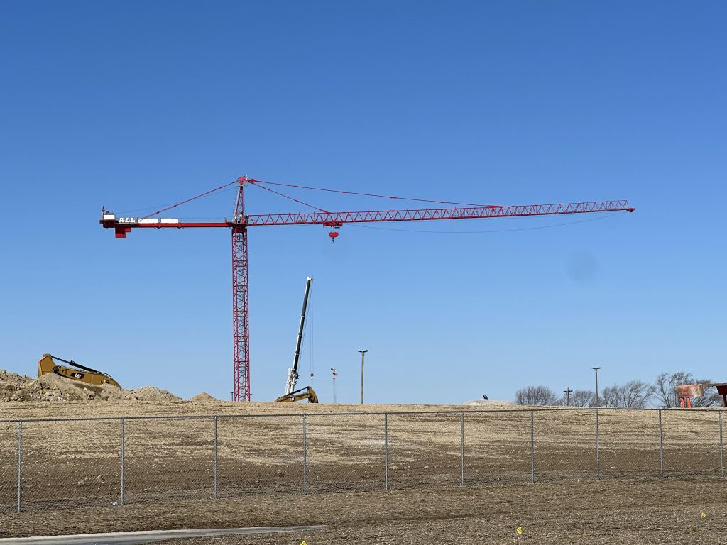 Red construction crane spans a dirt construction site with a chain-link fence in the foreground and a clear blue sky.