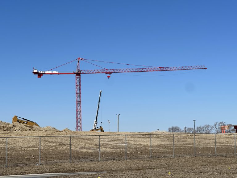 Red construction crane spans a dirt construction site with a chain-link fence in the foreground and a clear blue sky.