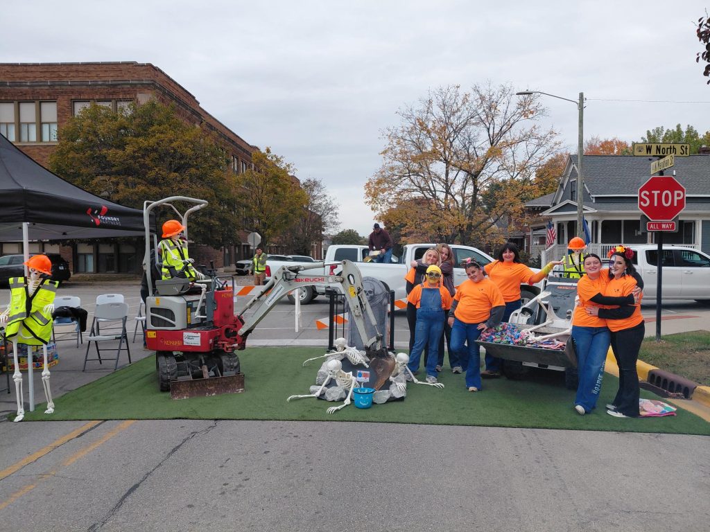 People in bright orange shirts pose with Halloween skeleton props on a city street, near construction equipment and a tent.